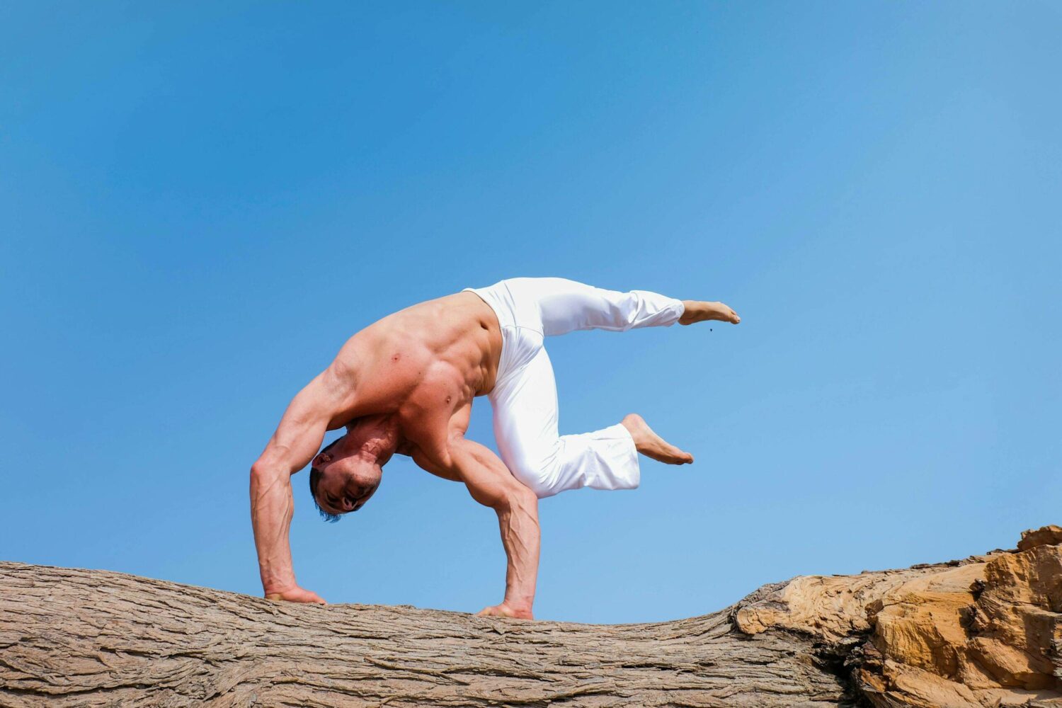 Mann führt anspruchsvolle Yoga-Pose auf einem Felsen unter blauem Himmel aus.